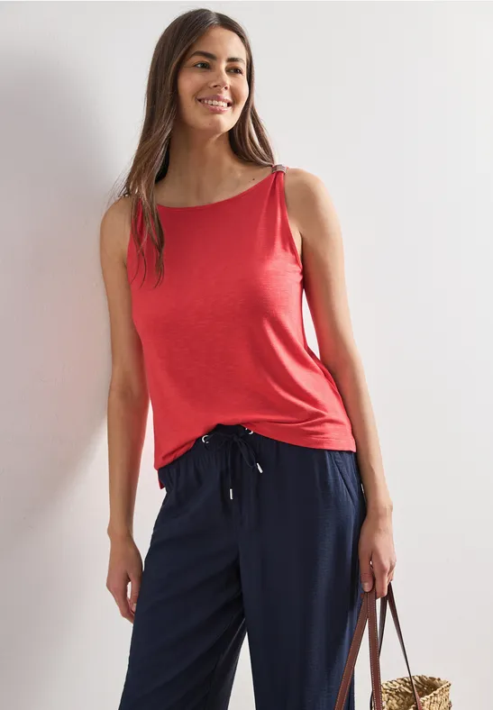 Vrouw in rood topje en donkerblauwe broek leunt nonchalant tegen een muur met een tas.