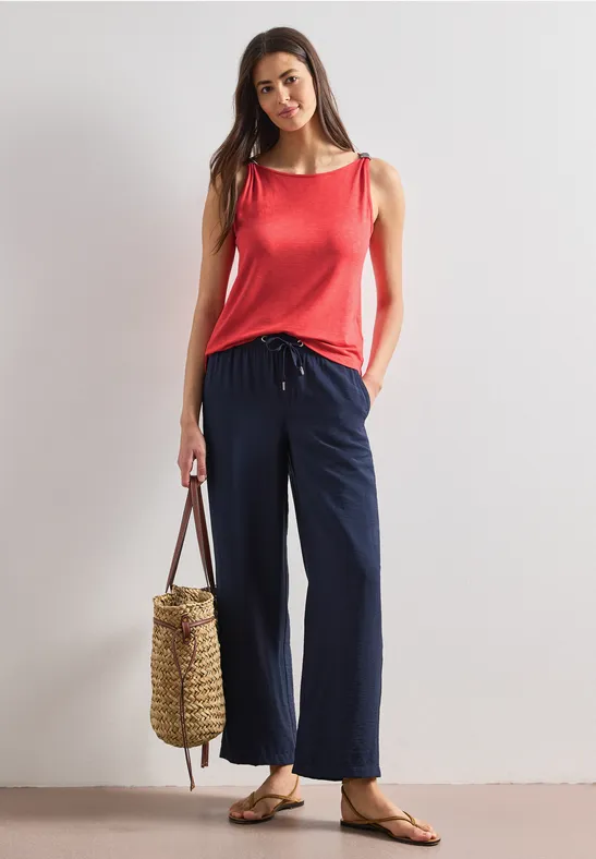 Vrouw in rood topje, wijde blauwe broek, sandalen en gevlochten tas tegen neutrale achtergrond.