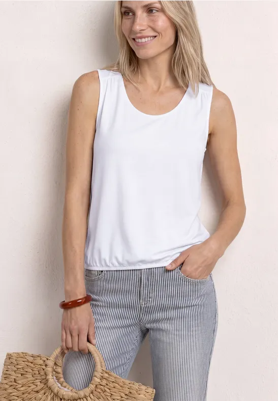 Vrouw in witte mouwloze top en grijze jeans met strooien tas.