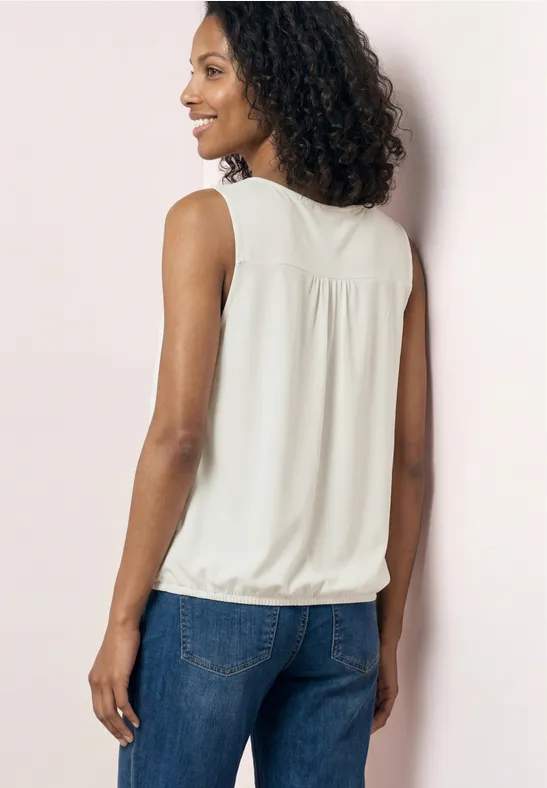 Eine Frau von hinten in weißer ärmelloser Bluse und blauen Jeans vor hellrosa Wand.