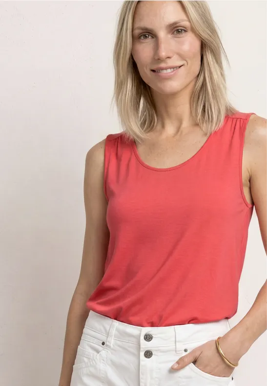 Vrouw in een rood tanktop en witte broek voor een neutrale achtergrond.
