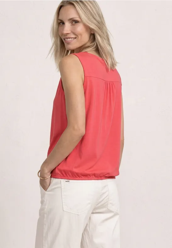 Vrouw van achteren in een koraalkleurige mouwloze top en witte broek, handen in de zakken.
