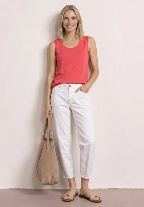 Een vrouw leunt tegen een muur, draagt een koraal top, witte broek, sandalen en een grote stoffen tas.