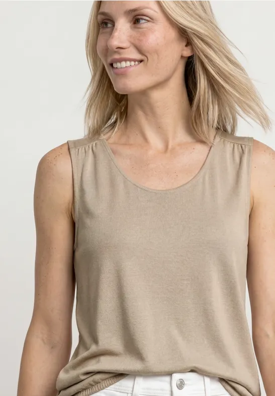 Glimlachende vrouw met blond haar in beige mouwloos shirt en witte broek.