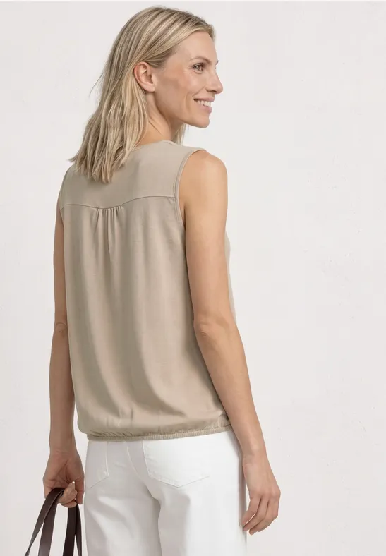 Vrouw met blond haar draagt beige mouwloze top en witte broek, rugaanzicht.