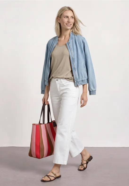 Vrouw in lichtblauw jasje, beige top, witte broek, sandalen en gestreepte tas, lopend voor neutrale achtergrond.