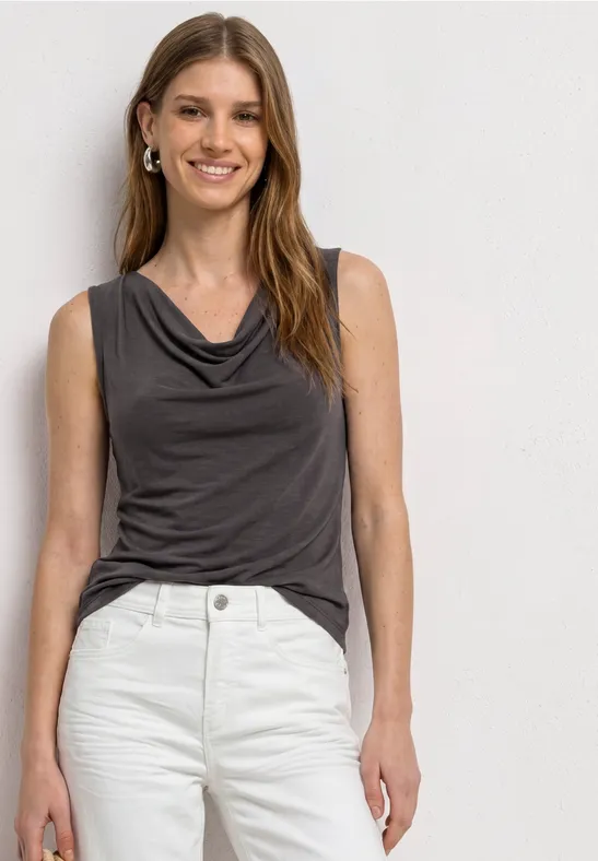 Vrouw met grijs mouwloos topje met watervalhals en witte spijkerbroek, voor een neutrale achtergrond.