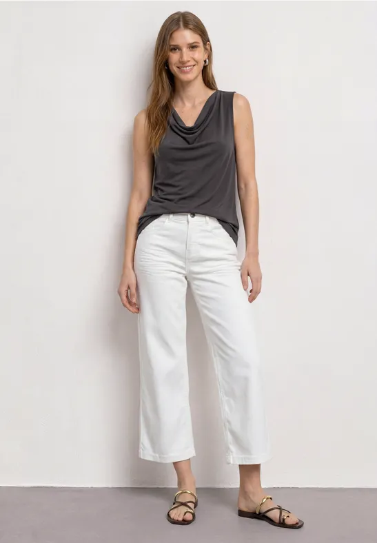Vrouw in mouwloze donkergrijze top, witte wijde broek en sandalen tegen een neutrale achtergrond.