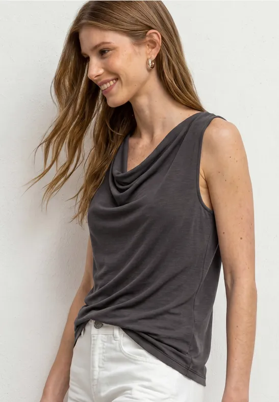 Vrouw met mouwloos, losvallend donkergrijs shirt en witte broek, met golvend haar en gouden oorbellen.
