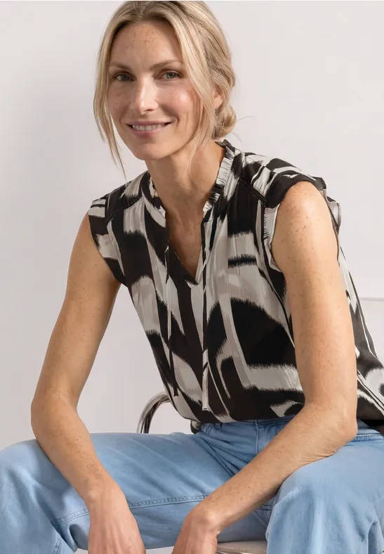 Een vrouw zit op een stoel, draagt een zwart-wit gemusterd mouwloos topje en een blauwe broek.