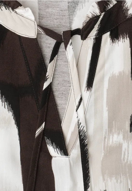 Close-up van een kledingstuk met abstract penseelstrekenpatroon in wit, bruin en zwart en V-hals met strik