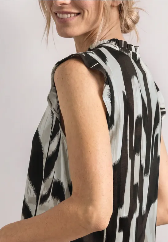 Een glimlachende vrouw draagt een mouwloos topje met een zwart-wit abstract patroon en kijkt over haar schouder.