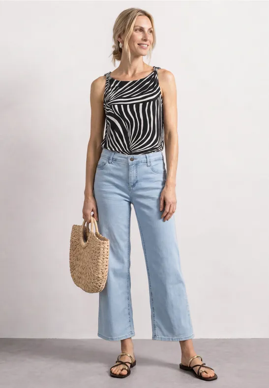 Eine Frau trägt ein gestreiftes Tanktop, helle Schlagjeans, schwarze Sandalen und hält eine geflochtene Strohtasche.