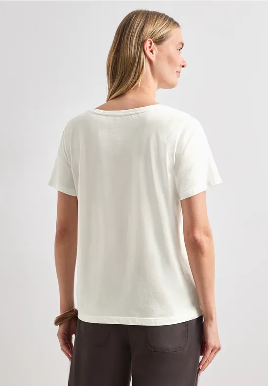 Eine Person steht mit dem Rücken zur Kamera, trägt ein weißes T-Shirt und eine dunkle, lockere Hose.