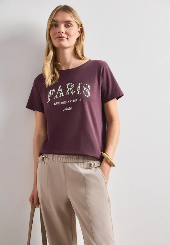 Vrouw draagt violet T-shirt met tekst en lichte broek, staat rechtop en houdt een tas vast.