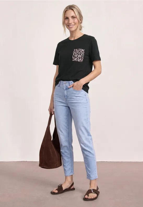 Vrouw met zwart t-shirt met patroon op borstzak, lichtblauwe jeans, zwarte sandalen en een bruine tas.