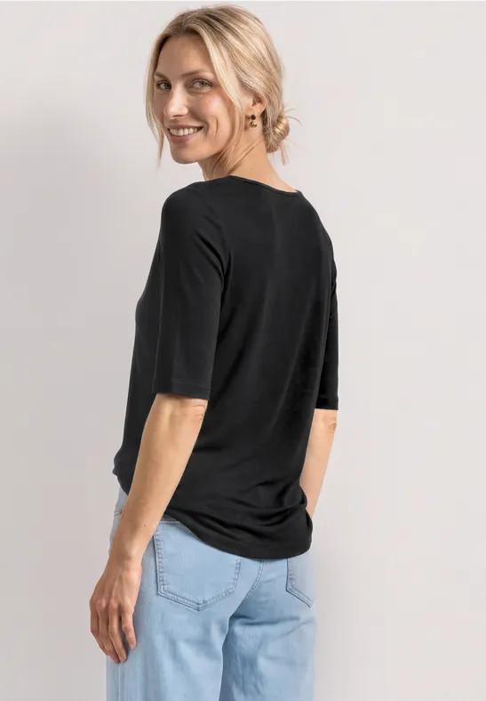 Frau von hinten in schwarzem Shirt mit halblangen Ärmeln und hellblauen Jeans vor neutralem Hintergrund.