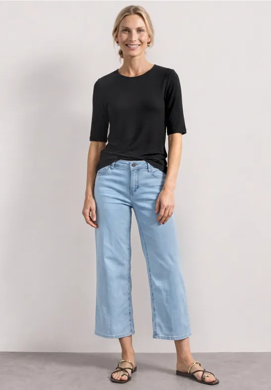 Frau in schwarzem Oberteil, hellblauen, knöchellangen Jeans und schwarzen Sandalen vor hellem Hintergrund.