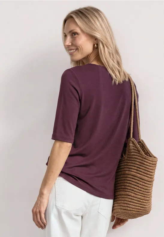 Vrouw met paarse T-shirt, witte broek en gevlochten tas, rug naar de camera en zijwaarts kijkend.