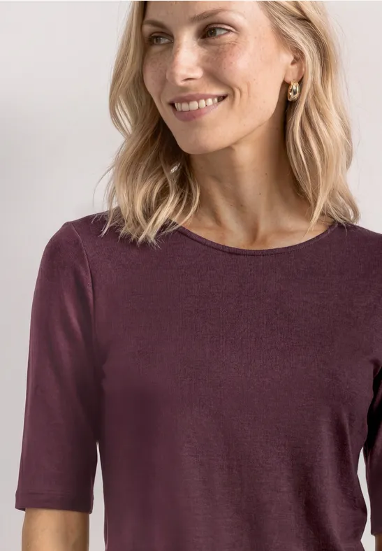 Een blonde vrouw glimlacht licht terwijl ze een bordeauxrood shirt met halve mouwen draagt voor een lichte achtergrond.