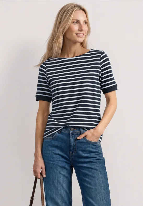 Vrouw in blauw-wit gestreept shirt, blond haar, spijkerbroek en tas, ontspannen houding.