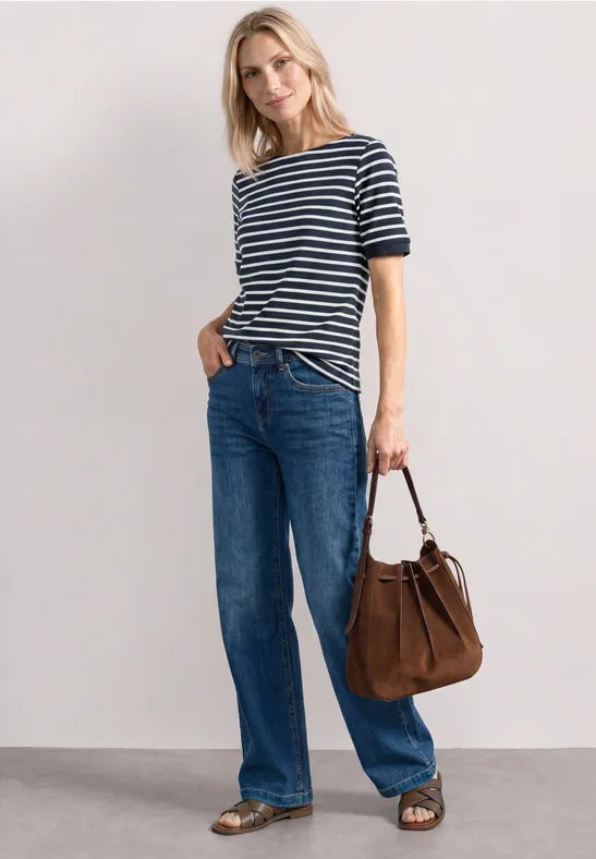 Vrouw in blauw-wit gestreepte trui, wijde jeans, bruine tas en sandalen tegen een neutrale achtergrond.