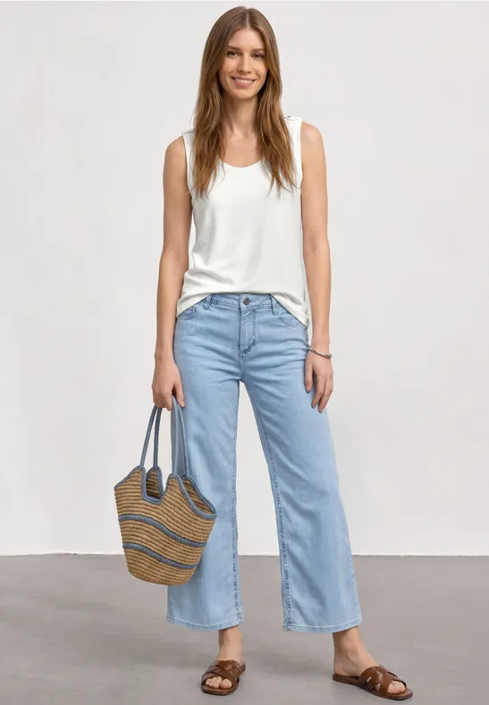 Frau trägt weiße Tanktop, helle Jeans, Sandalen und eine Strohtasche, stehend vor neutralem Hintergrund.
