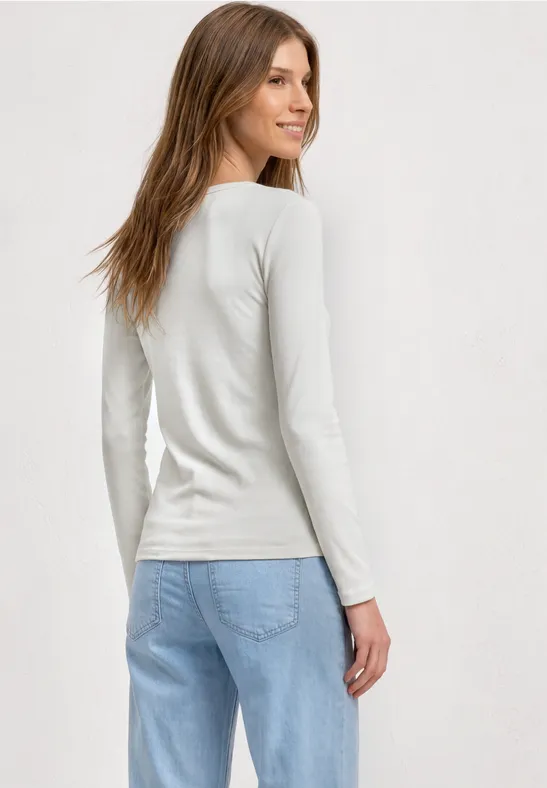 Femme en haut à manches longues blanc et jean bleu clair, regard par-dessus l'épaule, fond neutre.