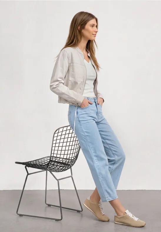 Femme appuyée sur chaise en métal, veste blanche, haut blanc, jean clair et baskets.