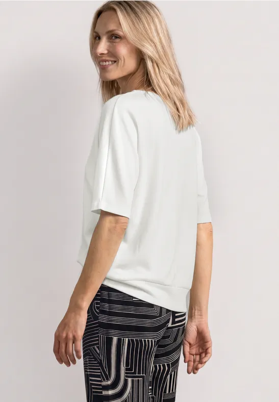 Femme de profil portant un t-shirt blanc et un pantalon noir à motifs sur fond clair.