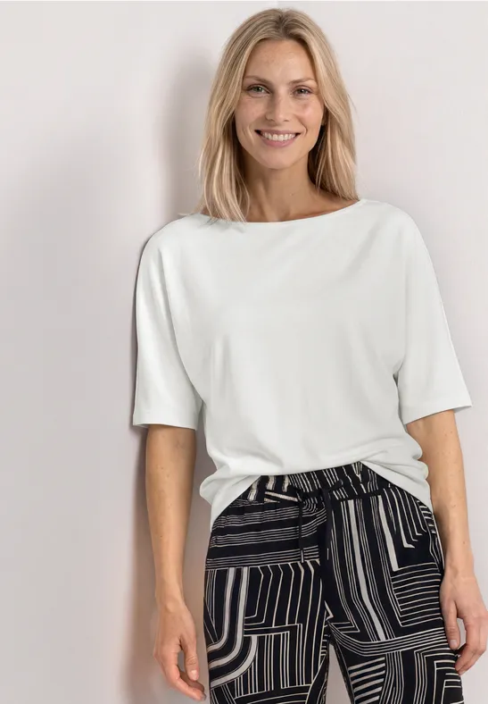 Femme en t-shirt blanc et pantalon noir à motifs, posant devant un fond neutre.