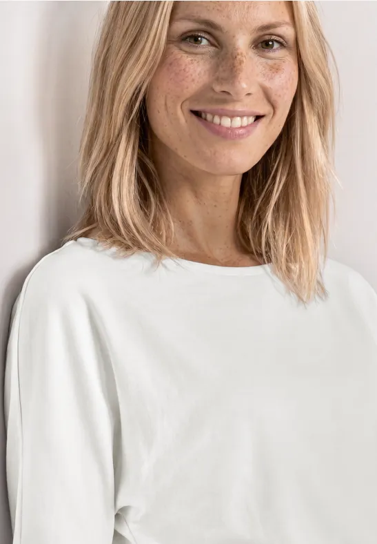 Femme blonde aux cheveux mi-longs portant un T-shirt blanc épuré devant un fond clair.