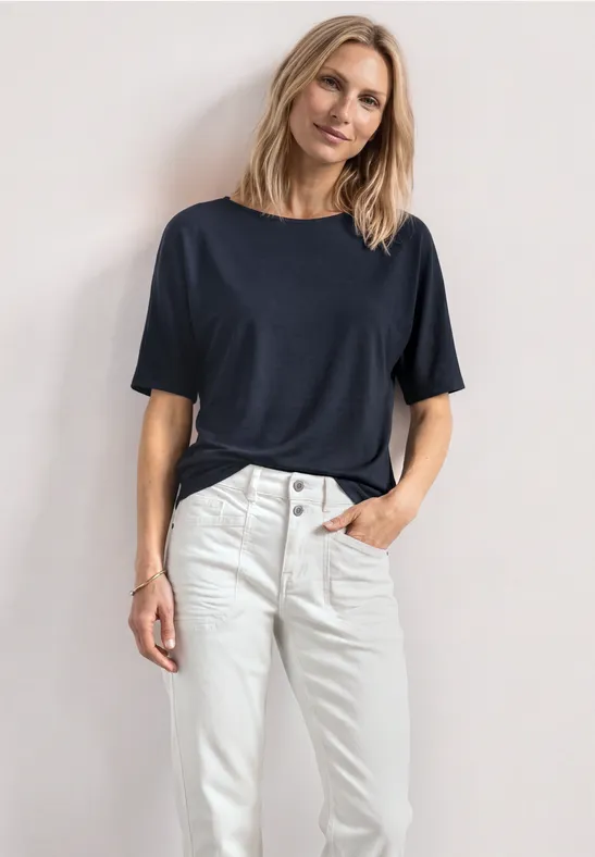 Femme portant un t-shirt bleu foncé et un jean blanc, debout de profil la main dans la poche.