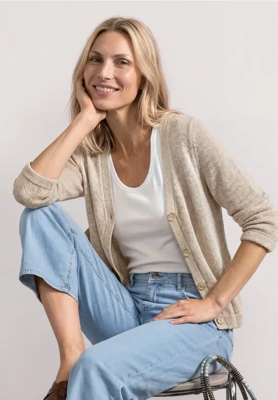 Une femme assise de façon décontractée porte un jean clair, un t-shirt blanc et un cardigan beige.