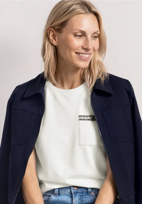 Frau mit blonderem Haar, trägt ein weißes T-Shirt, blaue Jeans, dunkle Jacke lässig über den Schultern.