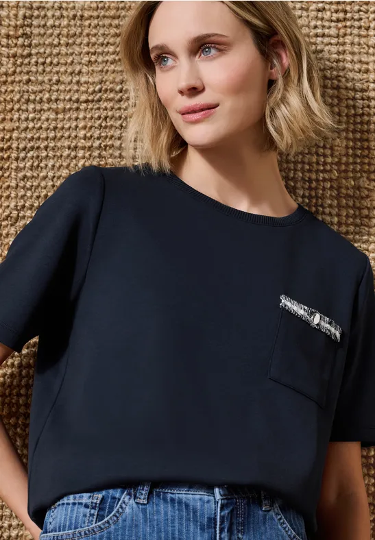 Une femme porte un top bleu marine à poche zippée, un jean, devant un fond en osier beige.
