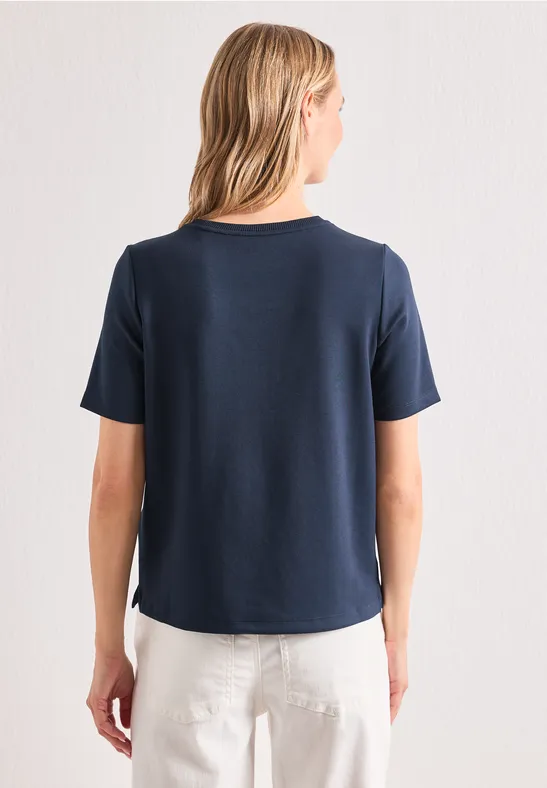 Femme de dos portant un T-shirt bleu marine et un pantalon blanc sur fond clair.