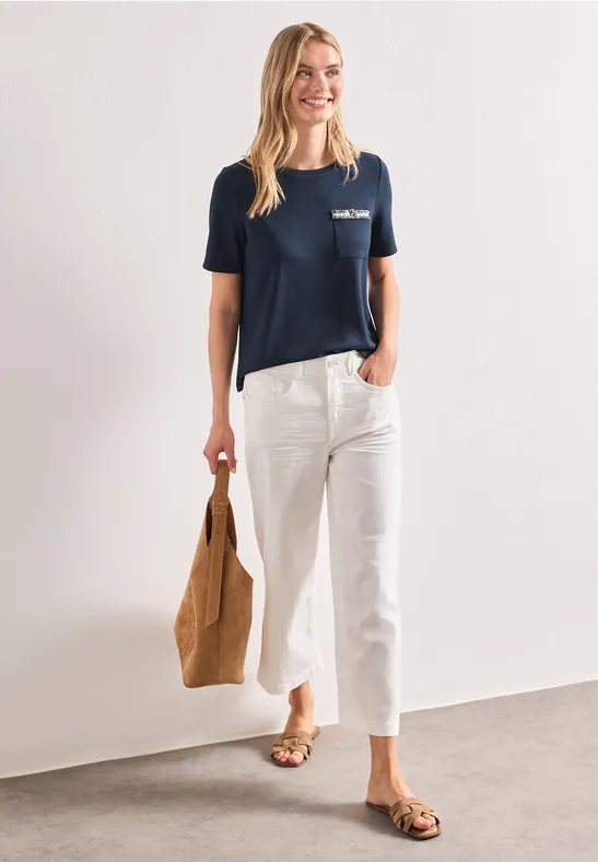Femme en t-shirt bleu marine, pantalon blanc court, sac marron et sandales sur fond blanc.