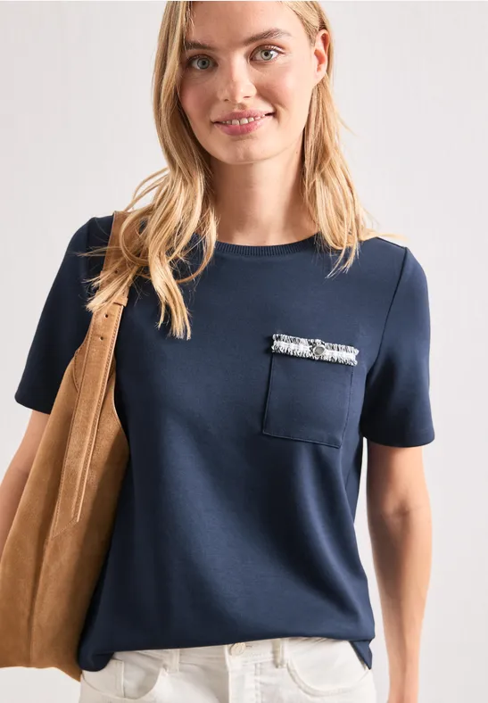 Femme portant un T-shirt bleu marine à manches courtes, poche ornée de paillettes, pantalon blanc et sac en suède.