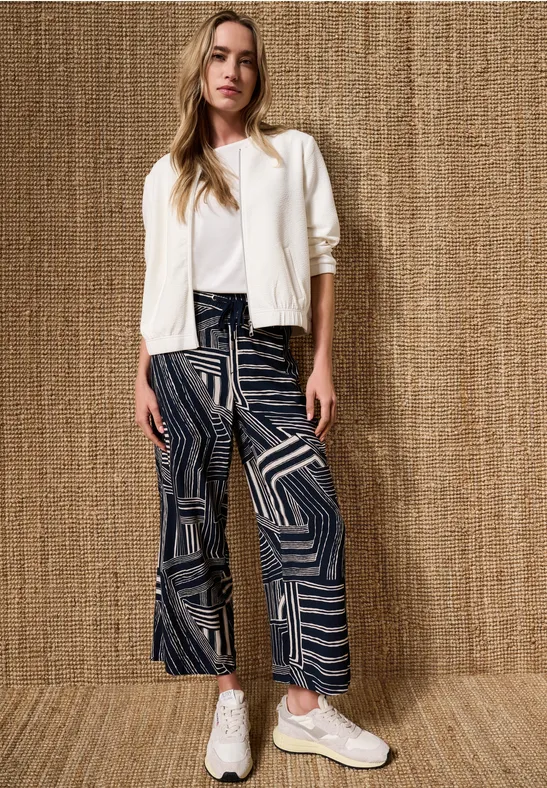 Femme en veste blanche, top noir, pantalon large à motifs et baskets devant un mur tressé.