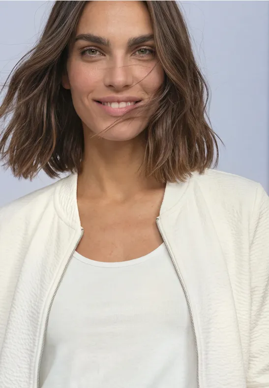 Femme aux cheveux châtains mi-longs souriante, portant un top blanc et un gilet clair sur fond neutre.