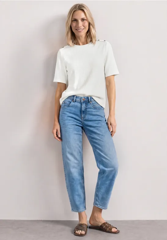 Femme en t-shirt blanc, jean bleu décontracté et sandales marron devant un fond neutre.
