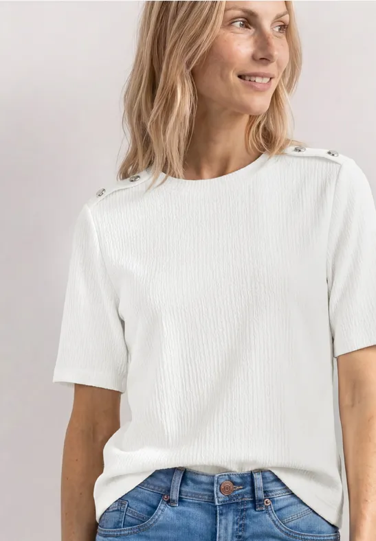 Femme en t-shirt blanc à manches courtes avec boutons aux épaules et jean bleu, torse visible, visage flou.