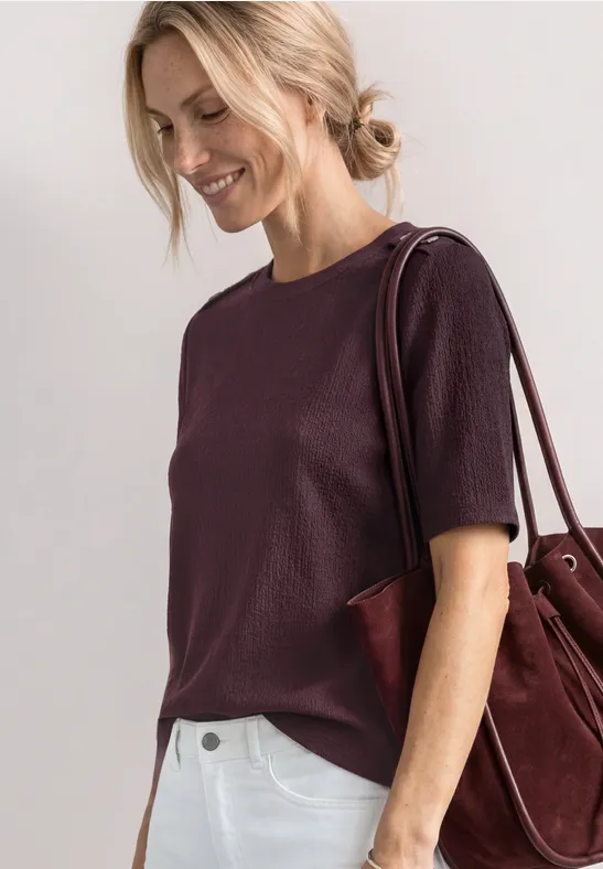 Vrouw met violet T-shirt met korte mouwen en een bordeaux schoudertas tegen een lichte achtergrond.