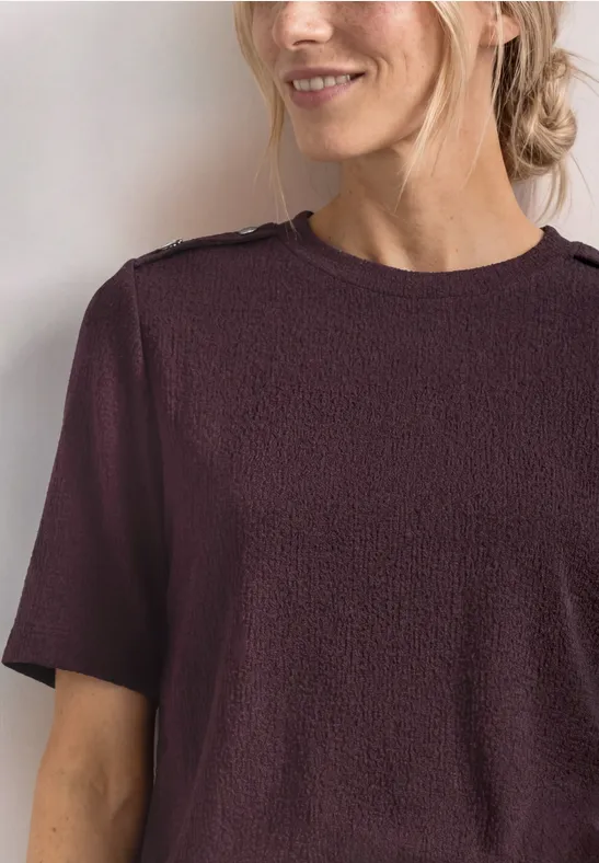 Close-up van een glimlachende vrouw met opgestoken blond haar en een bordeauxkleurig kort T-shirt.