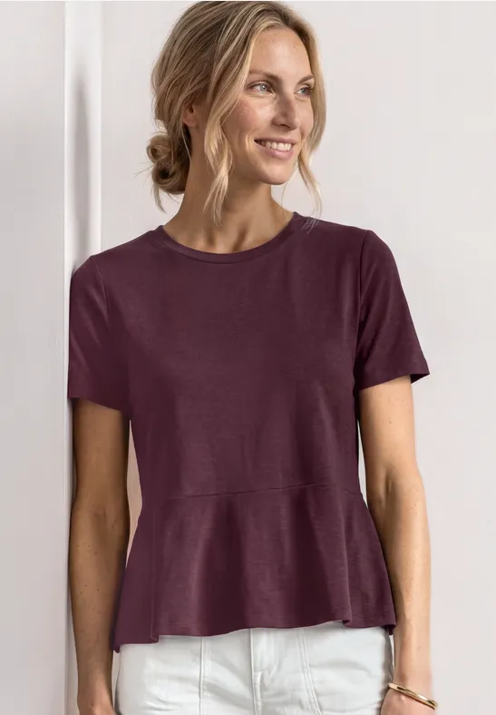 Femme portant un t-shirt violet à manches courtes avec basque évasée et pantalon blanc sur fond neutre.