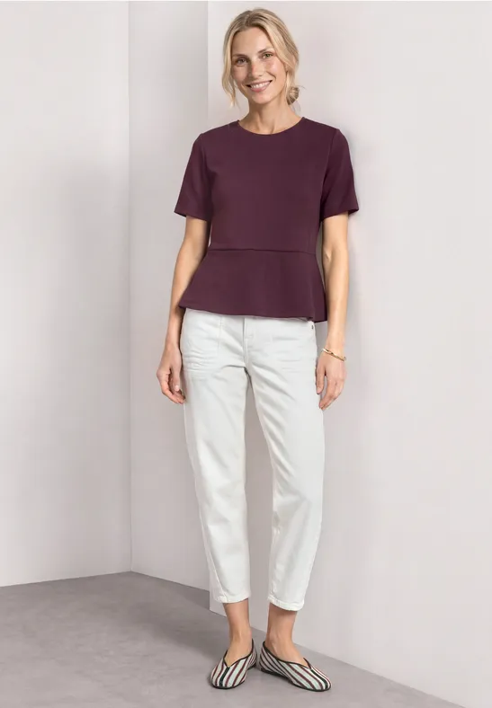 Femme portant un t-shirt violet, un pantalon blanc et des ballerines rayées sur fond neutre.