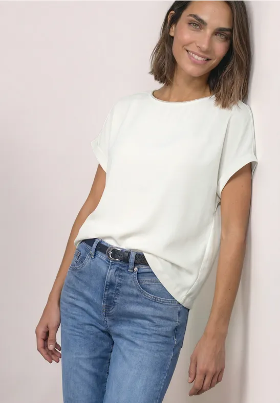 Lächelnde Frau trägt lockeres weißes T-Shirt und blaue Jeans vor hellem Hintergrund.