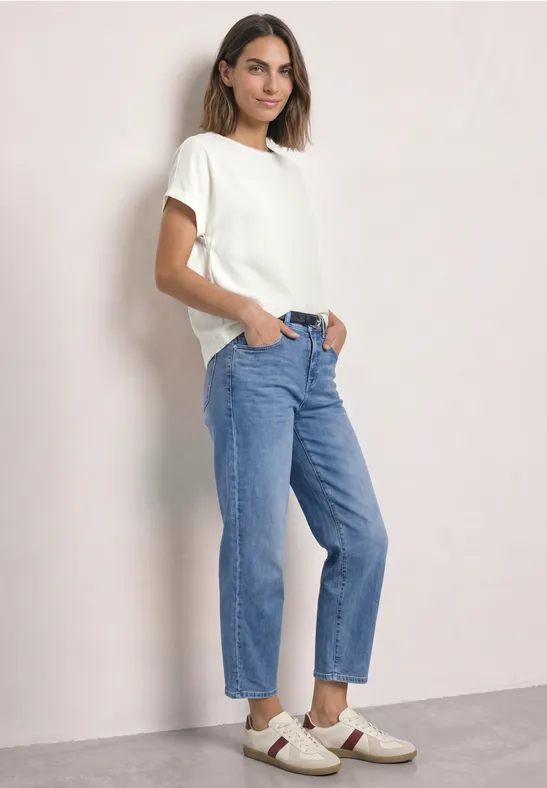 Eine junge Frau steht lässig vor hellem Hintergrund in weißem T-Shirt, blauen Jeans und weißen Schuhen.