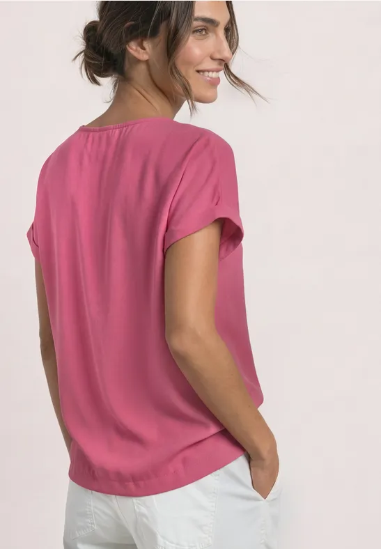 Eine seitliche Aufnahme einer lächelnden Frau in pinkem T-Shirt und weißen Hosen mit Händen in den Taschen.
